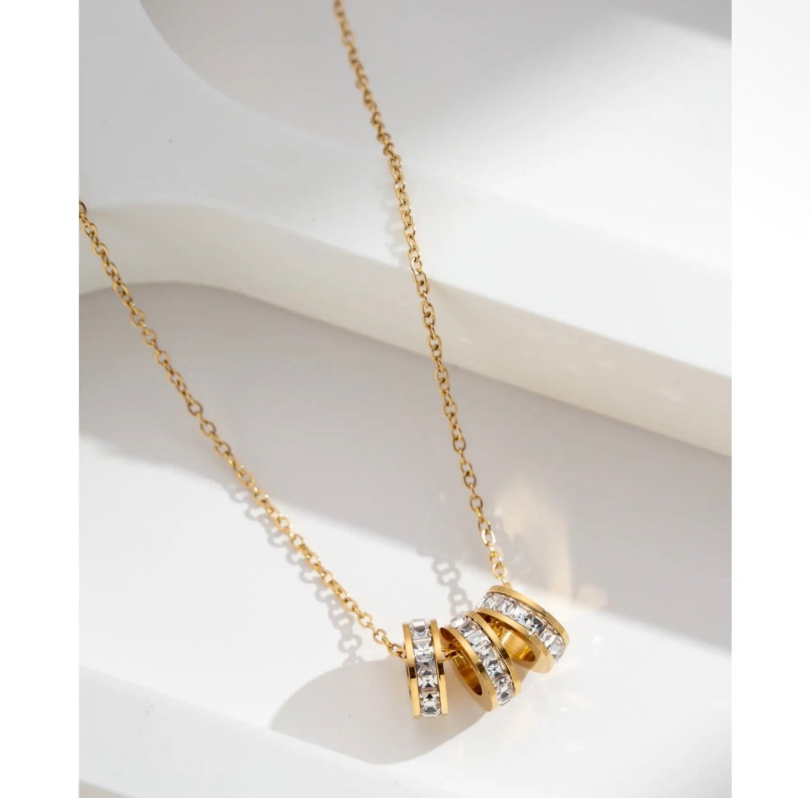 Collier pendentif triple anneaux dorés serties de zircons sur fond blanc